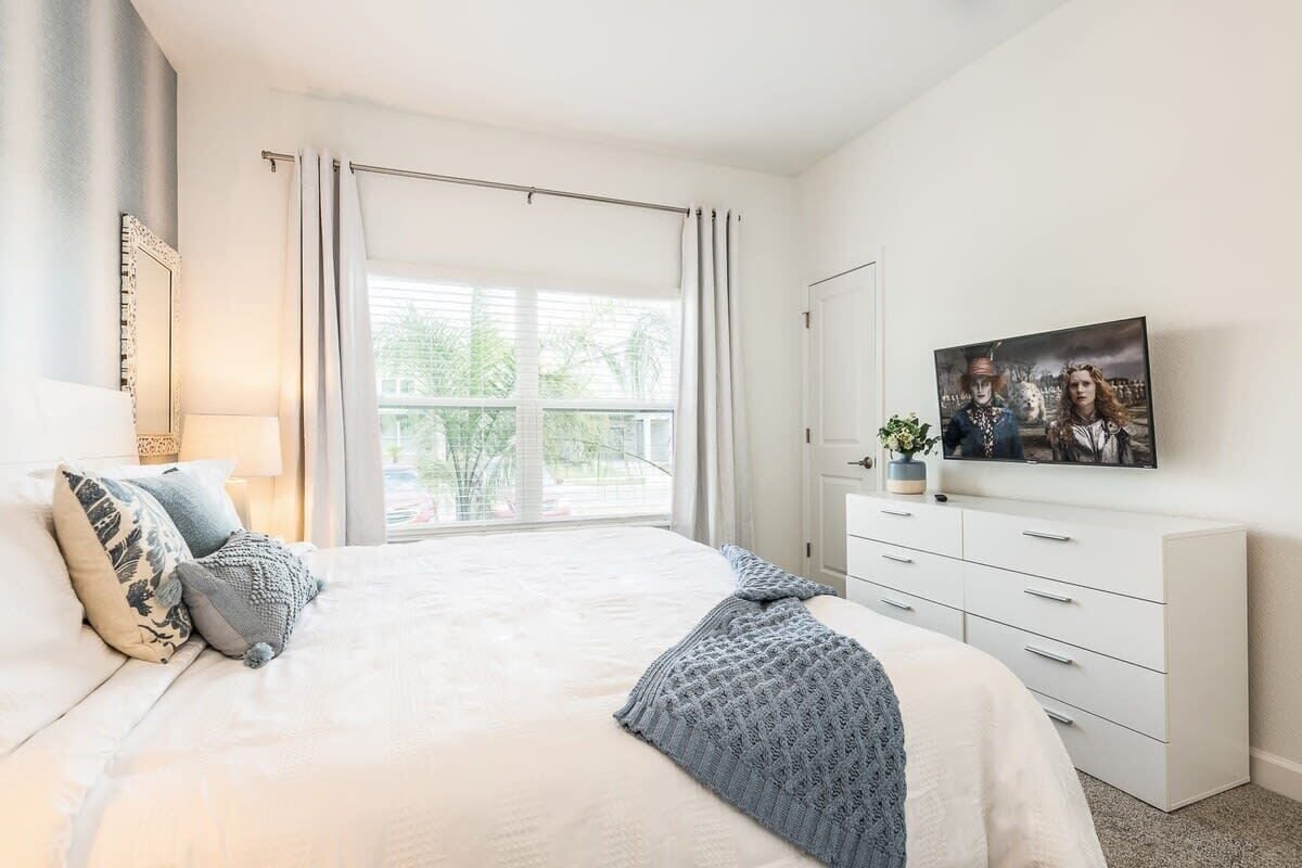 A bedroom with a bed, a TV, a dresser, and a window with curtains.