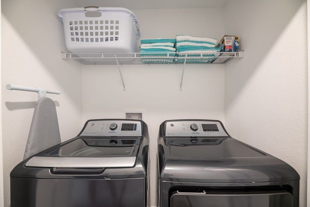 A laundry area with two front-loading washing machines, a white laundry basket, and a shelf with folded towels and a box.