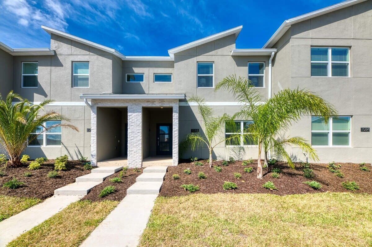 A modern two-story building with a flat roof, a central entrance with a stone accent, and a landscaped front yard with palm trees and shrubs.