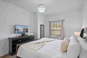 A bedroom with a bed, a dresser, a television, a window with curtains, and a chandelier. - Modern 3 Bed Villa, Newly Renovated, Prime Location (Kissimmee)