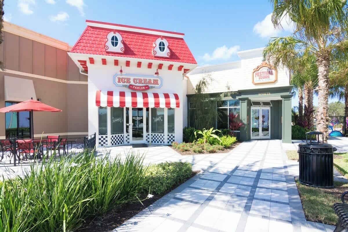 An ice cream parlor with a red roof and striped awning, located in a plaza with a tiled walkway and outdoor seating.