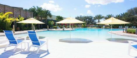 A pool area with lounge chairs and umbrellas, surrounded by palm trees and a brick building.
