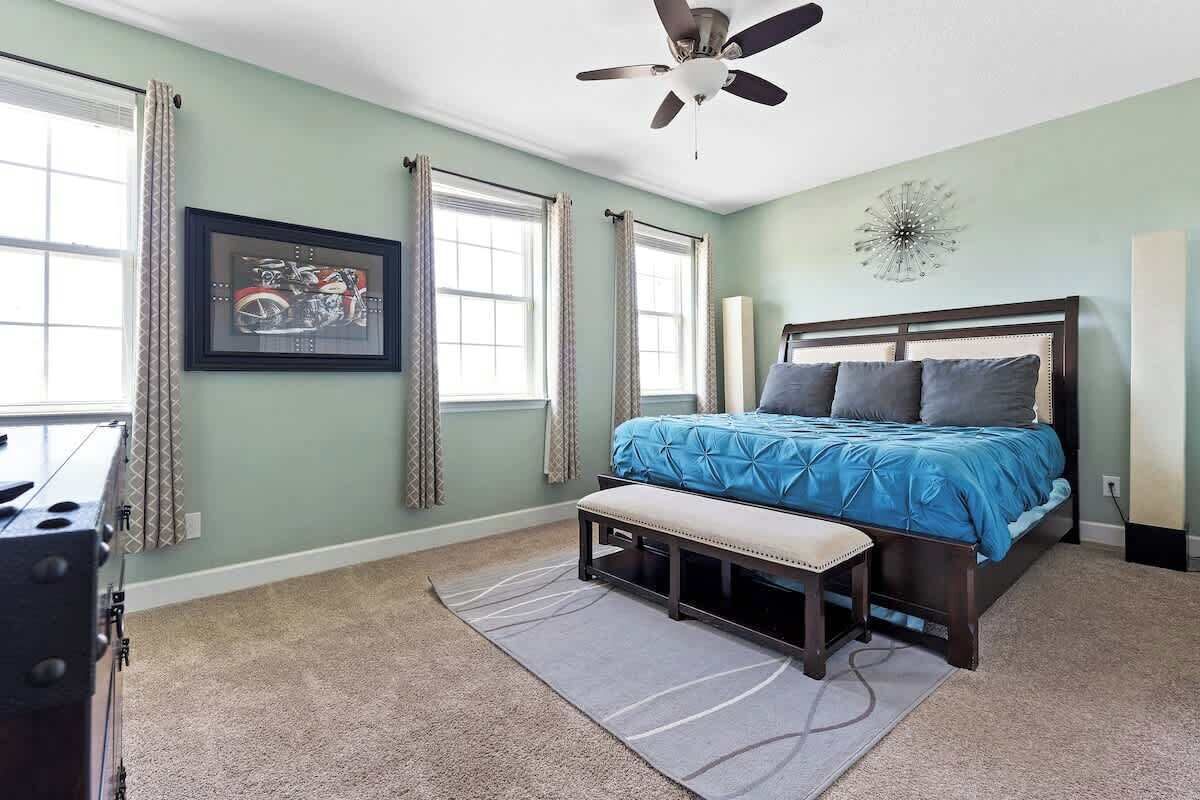 Une chambre à coucher avec un lit, un ventilateur de plafond, un tableau encadré au mur et une fenêtre avec des rideaux.