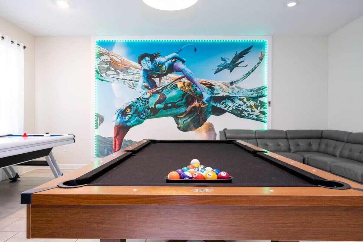 A pool table with a colorful ball rack in front, a sofa, and a mural with a flying character on the wall.