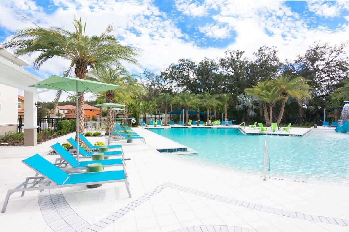 A swimming pool area with lounge chairs and umbrellas, surrounded by palm trees and a clear sky.
