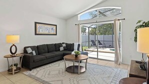 Ein modernes Wohnzimmer mit einer Eckcouch, einem runden Couchtisch, einer Lampe und einem großen Fenster mit Vorhängen.