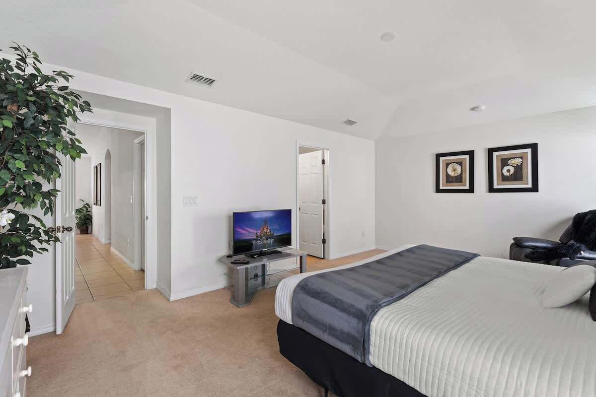 Un dormitorio moderno con una cama, un televisor y un escritorio. Hay una puerta que da a otra habitación y una planta en la esquina.