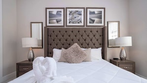 Une chambre d’hôtel avec un grand lit, deux tables de chevet, une tête de lit capitonnée et trois tableaux encadrés accrochés au mur.