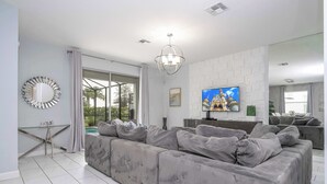 A modern living room with a grey sectional sofa, a glass coffee table, a TV, and a large mirror. - Home 6bed , Themed Rooms, Game Room, Storey Lake (Kissimmee)