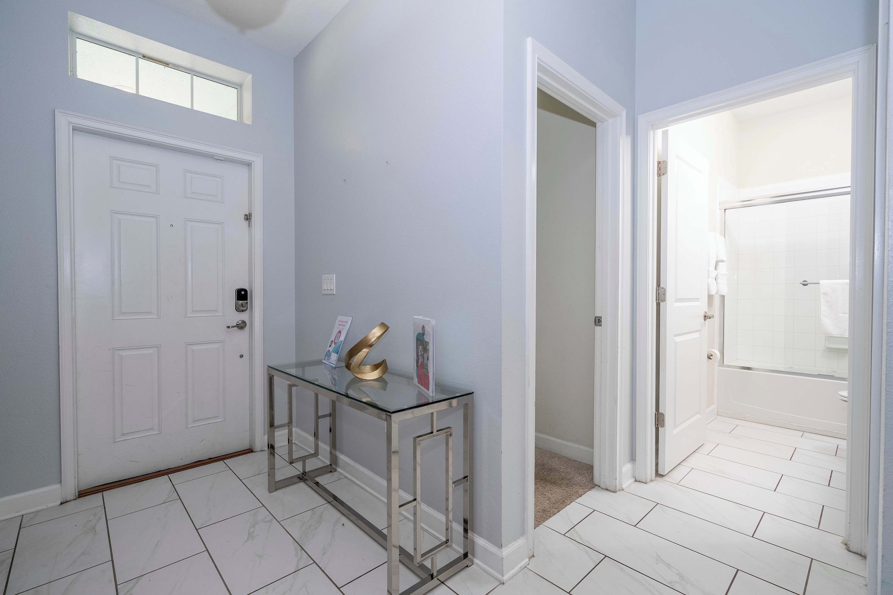Un pasillo con una puerta blanca, una mesita de consola de vidrio con objetos, y un baño visible a través de una puerta abierta.