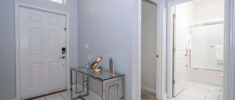Un pasillo con una puerta blanca, una mesita de consola de vidrio con objetos, y un baño visible a través de una puerta abierta.