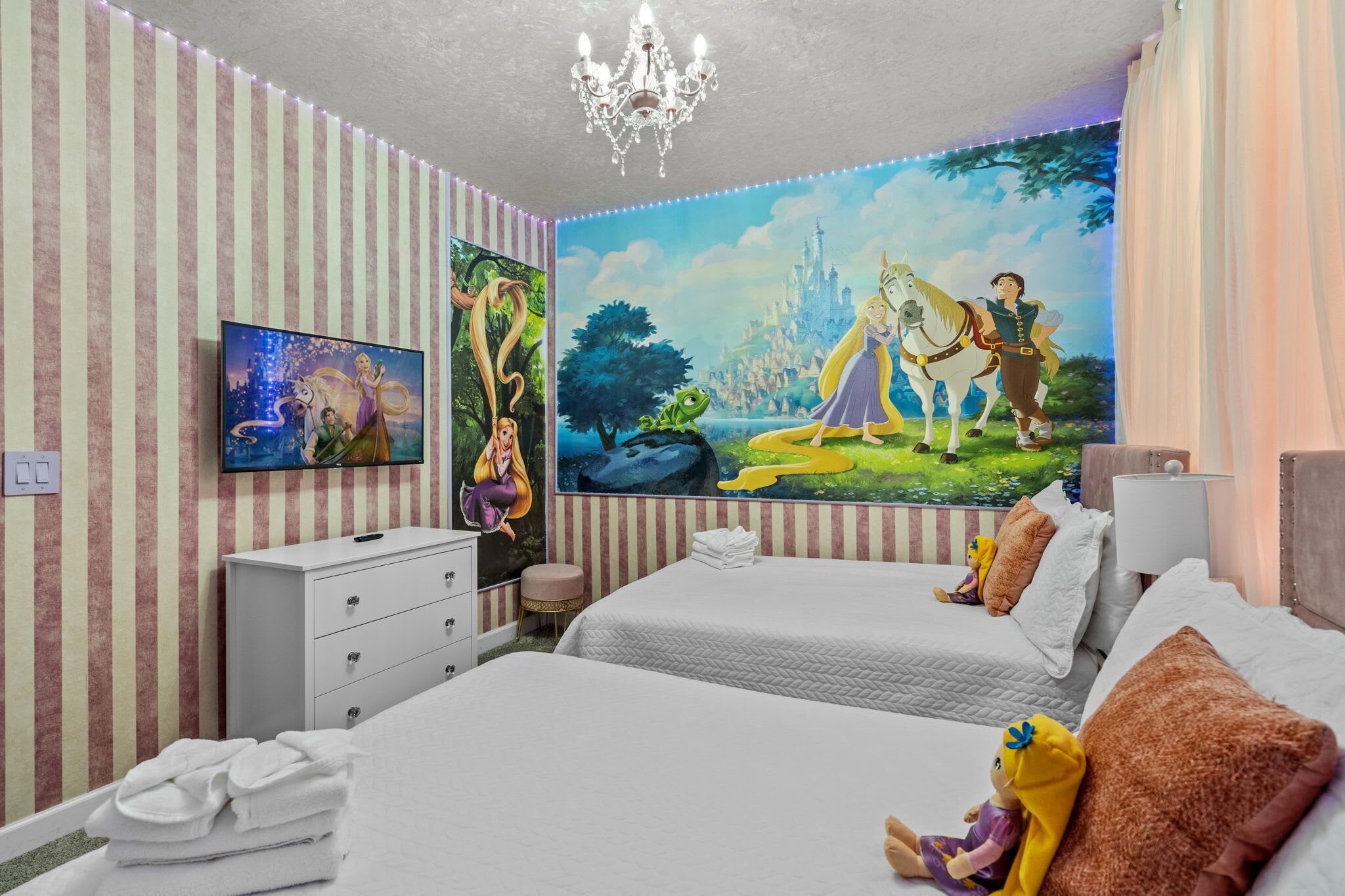A hotel room with striped wallpaper, a chandelier, a mural, a TV, a white dresser, two beds, and plush toys.