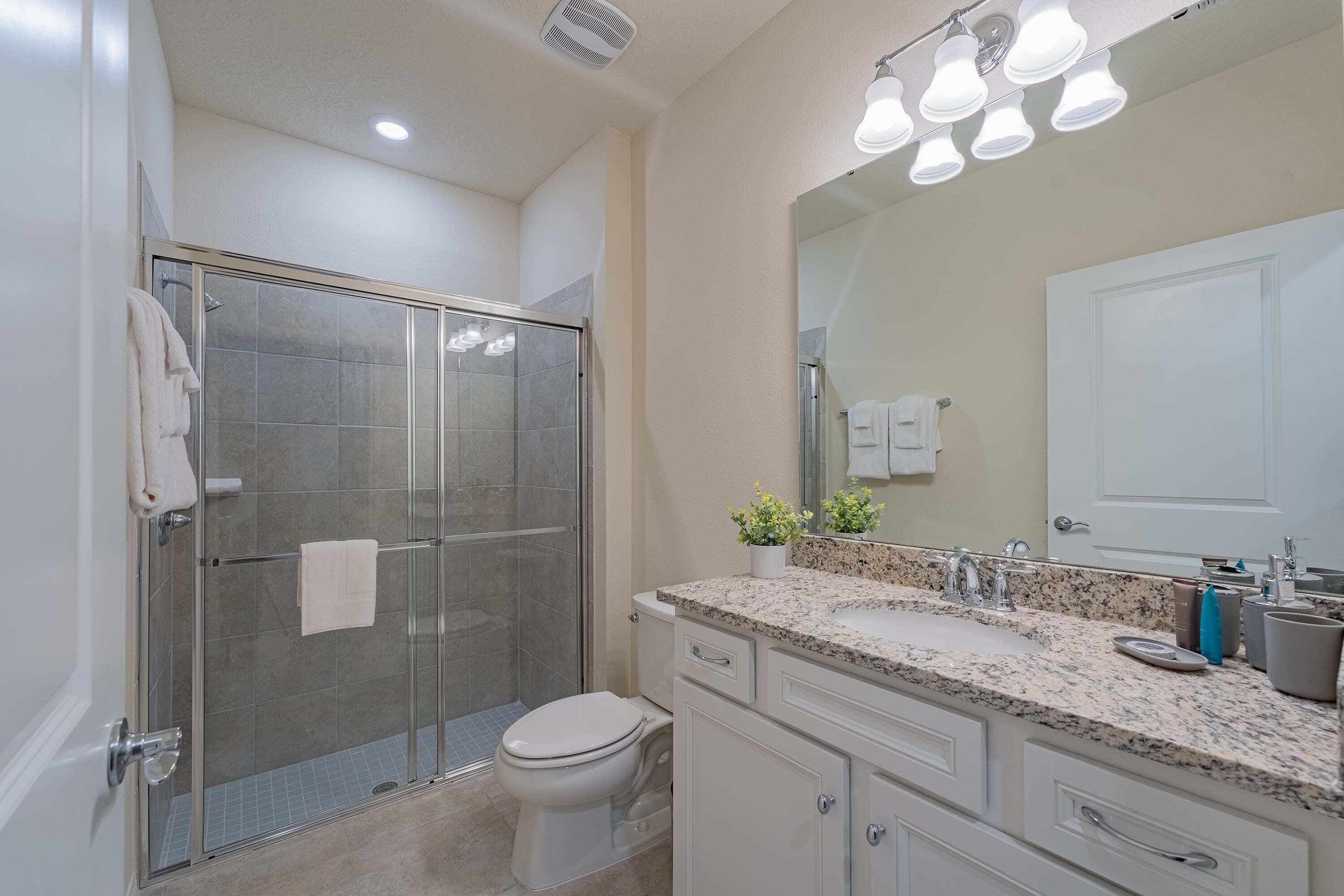 A modern bathroom with a glass-enclosed shower, a white toilet, a granite countertop, and a large mirror.