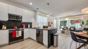 Una cocina moderna con microondas, horno y lavaplatos, un comedor con mesa y sillas, y vistas al exterior a través de amplias ventanas.