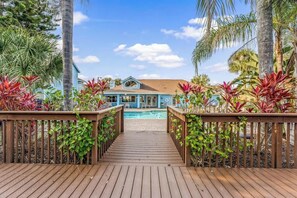 Eine Holzterrasse führt zu einem Haus mit Schwimmbecken, umgeben von tropischen Pflanzen.