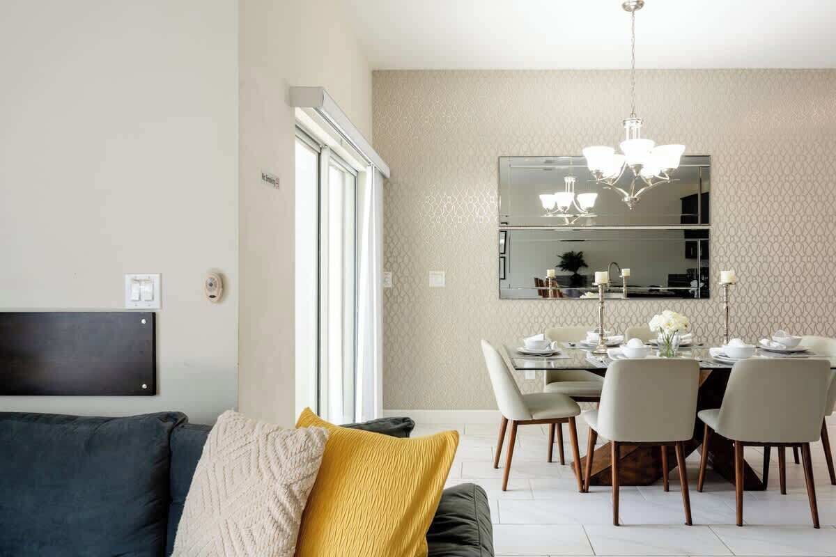 A modern living room with a sofa, a dining table with chairs, a chandelier, and a large mirror on the wall.
