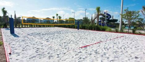 A sandy beach area with a volleyball net, palm trees, and a water slide structure in the background.