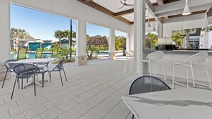 En udendørs patio med borde og stole, store vinduer samt udsigt til pool og bygninger.