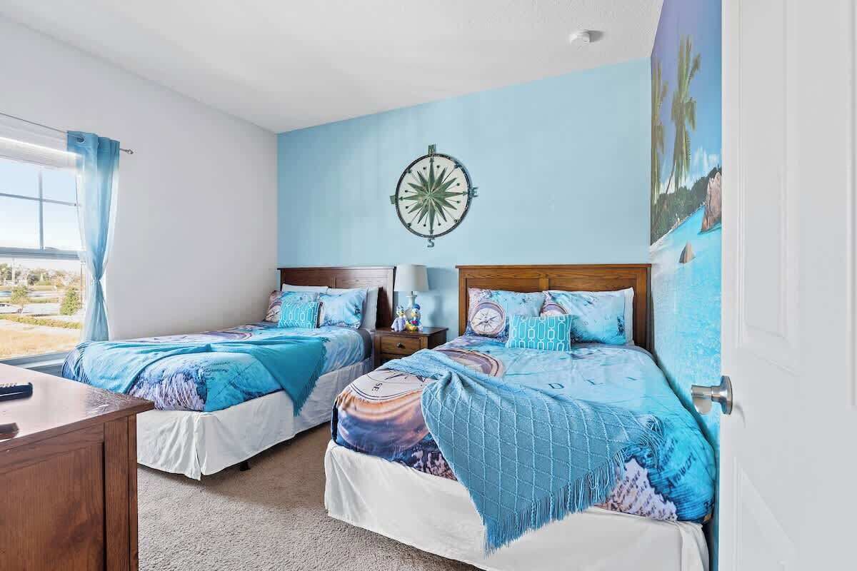 A bedroom with two beds, a wooden nightstand, a desk, a compass wall decoration, and a large window with curtains.