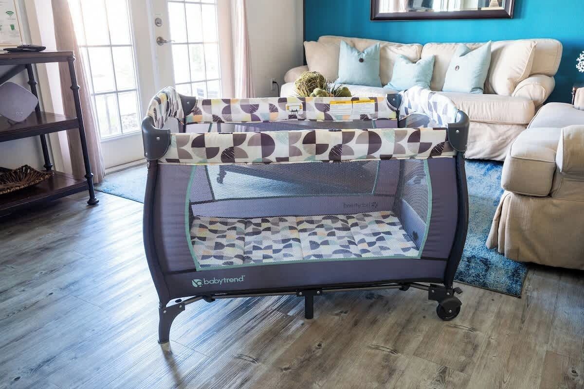 Un parc pour bébé au motif géométrique, placé dans une pièce dont le mur est bleu, qui contient un canapé beige et dont le sol est en bois.