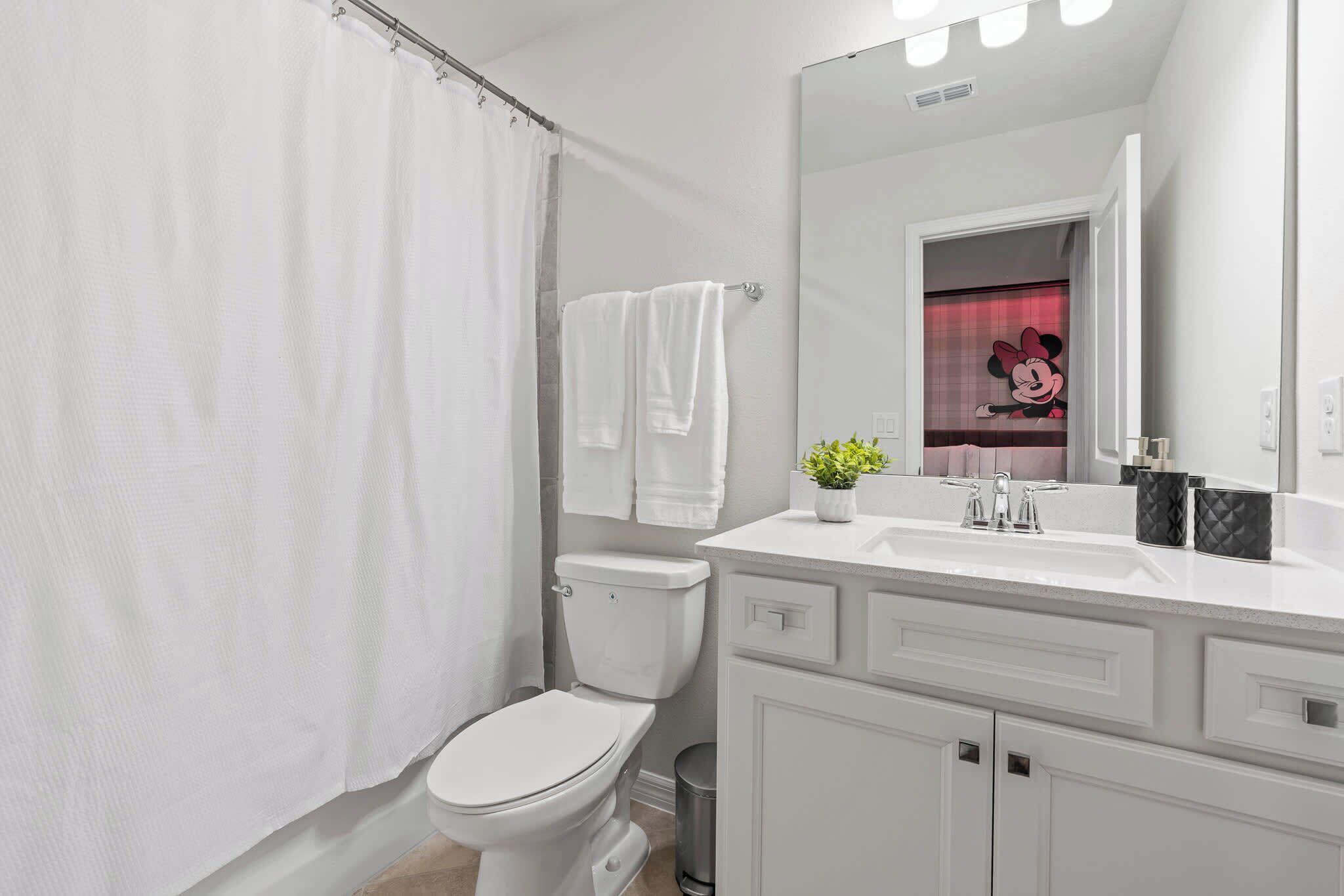 A bathroom with a white toilet, a shower curtain, a white sink with a mirror, and a Mickey Mouse decoration.