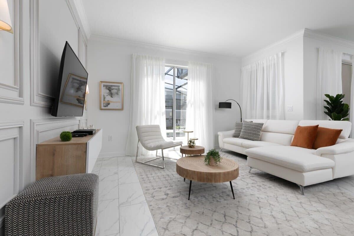 Ein modernes Wohnzimmer mit einem weißen Sofa, einem runden Couchtisch, einem weißen Sessel, einem an der Wand befestigten Fernseher und einem großen Fenster mit durchsichtigen Vorhängen.