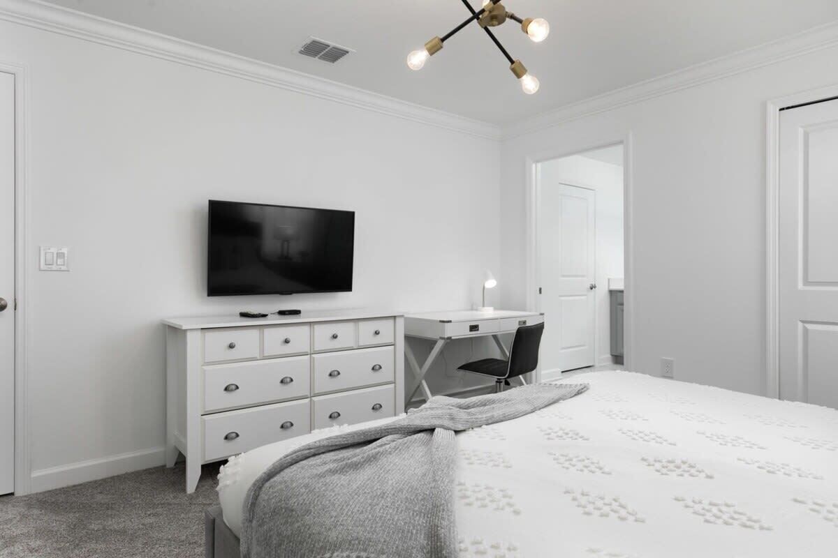 A bedroom with a bed, a dresser, a desk, a chair, a television, and a ceiling light fixture.
