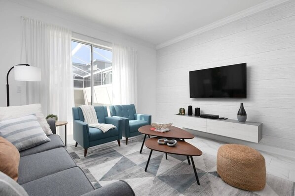 A modern living room with a sofa, armchair, coffee table, and ottoman. A flat-screen TV is mounted on the wall, and there is a window with curtains. - Chic Modern Home- Mickey & Star Wars Magic (Davenport)