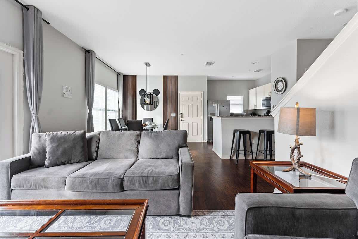 Een moderne woonkamer met een grijze bank, een salontafel en een lamp. Achtergrond is een keuken met een bargedeelte en krukken.