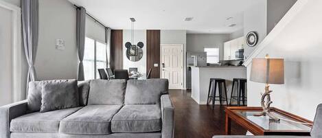 Een moderne woonkamer met een grijze bank, een salontafel en een lamp. Achtergrond is een keuken met een bargedeelte en krukken.