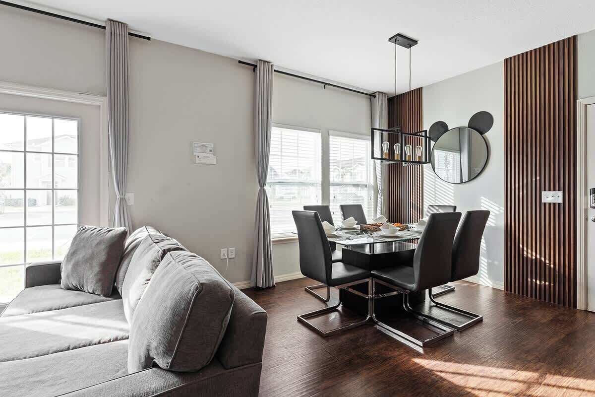 Un salon moderne avec un canapé gris, une table à manger avec des chaises, un lustre et un miroir mural orné d’un motif représentant un visage d’ours.