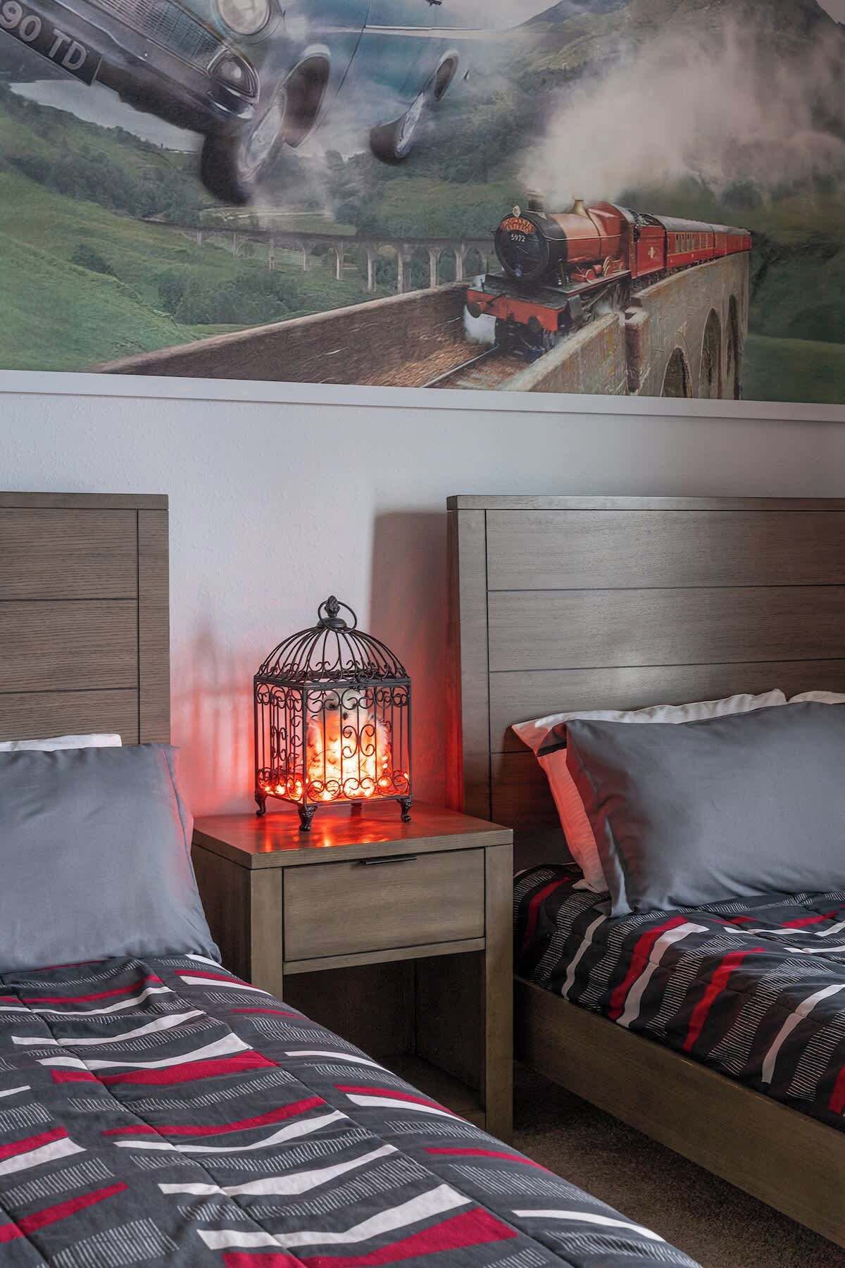 A bedroom with two beds, a nightstand with a lantern, and a mural of a train on the wall.