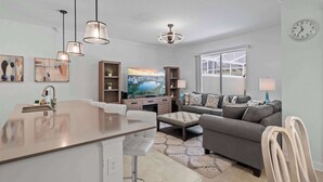 A modern living room with a kitchen area, a television, and a clock on the wall. - Magical Themed Home With Pool in Top Resort (Davenport)