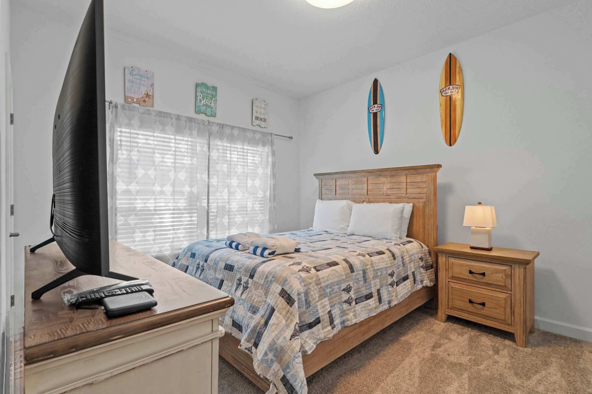 A bedroom with a bed, a nightstand with a lamp, a TV on a desk, and two surfboards on the wall.