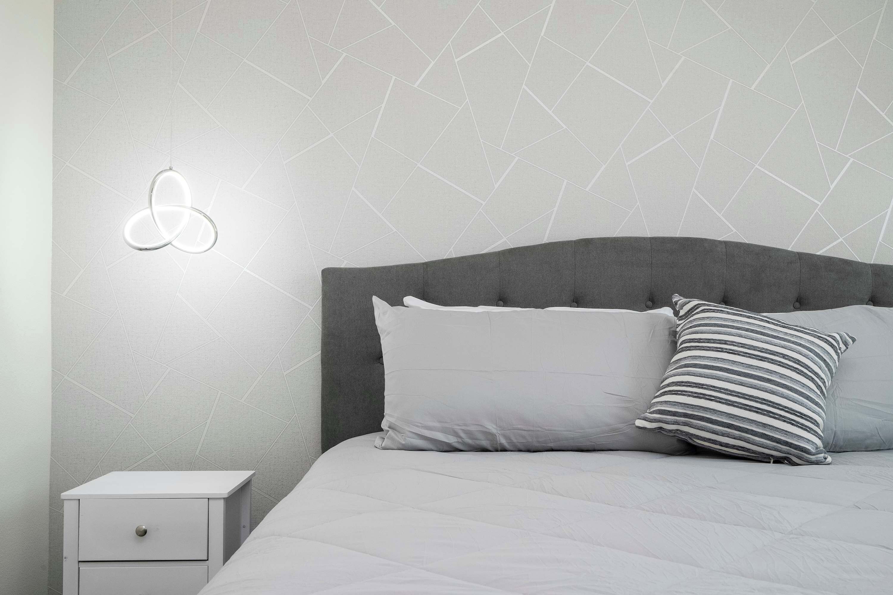 A bed with a grey headboard, a white bedside table with a drawer, and a wall-mounted light fixture.