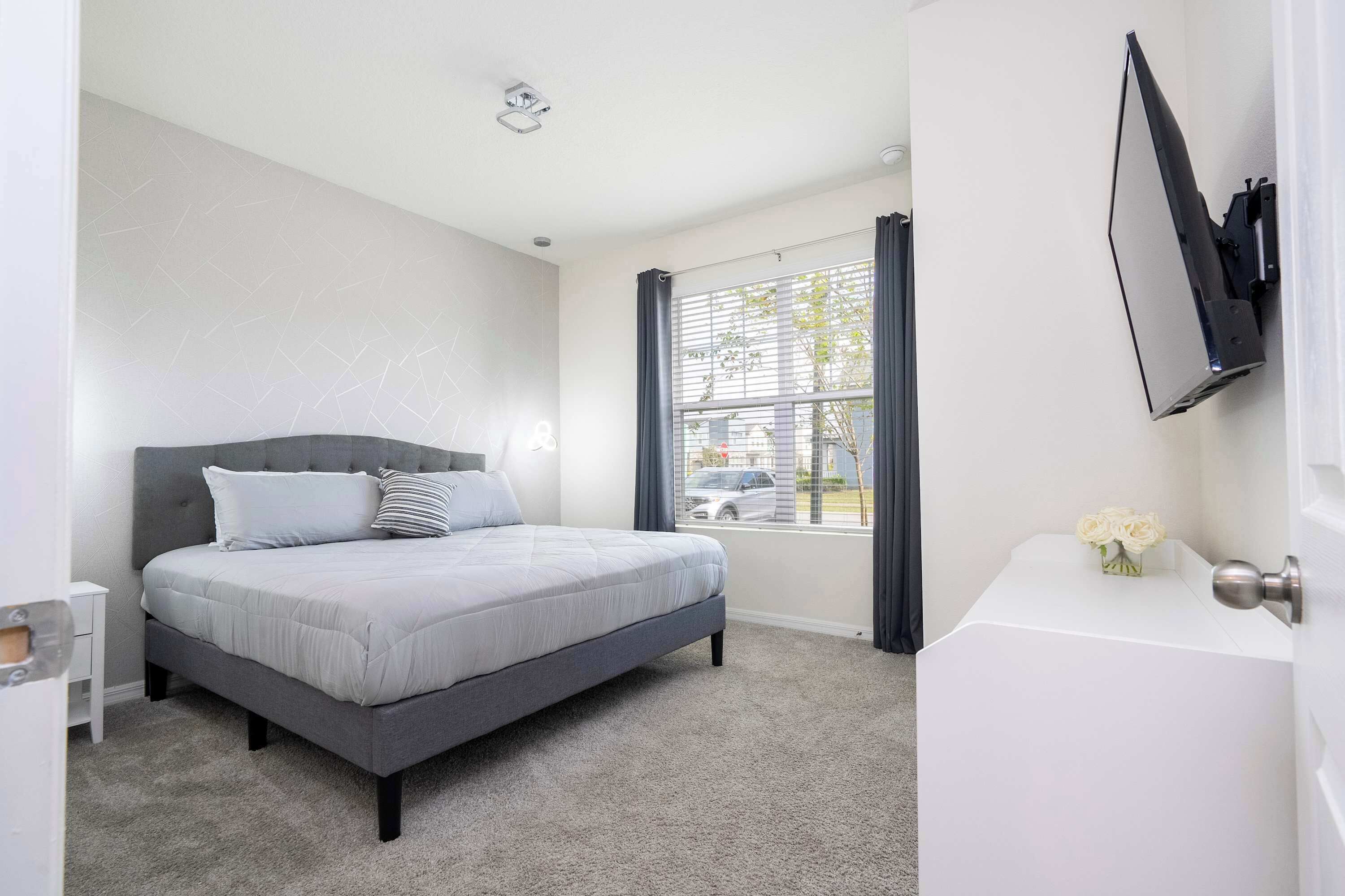A bedroom with a bed, a window with curtains, a flat-screen TV mounted on the wall, and a small bedside table.
