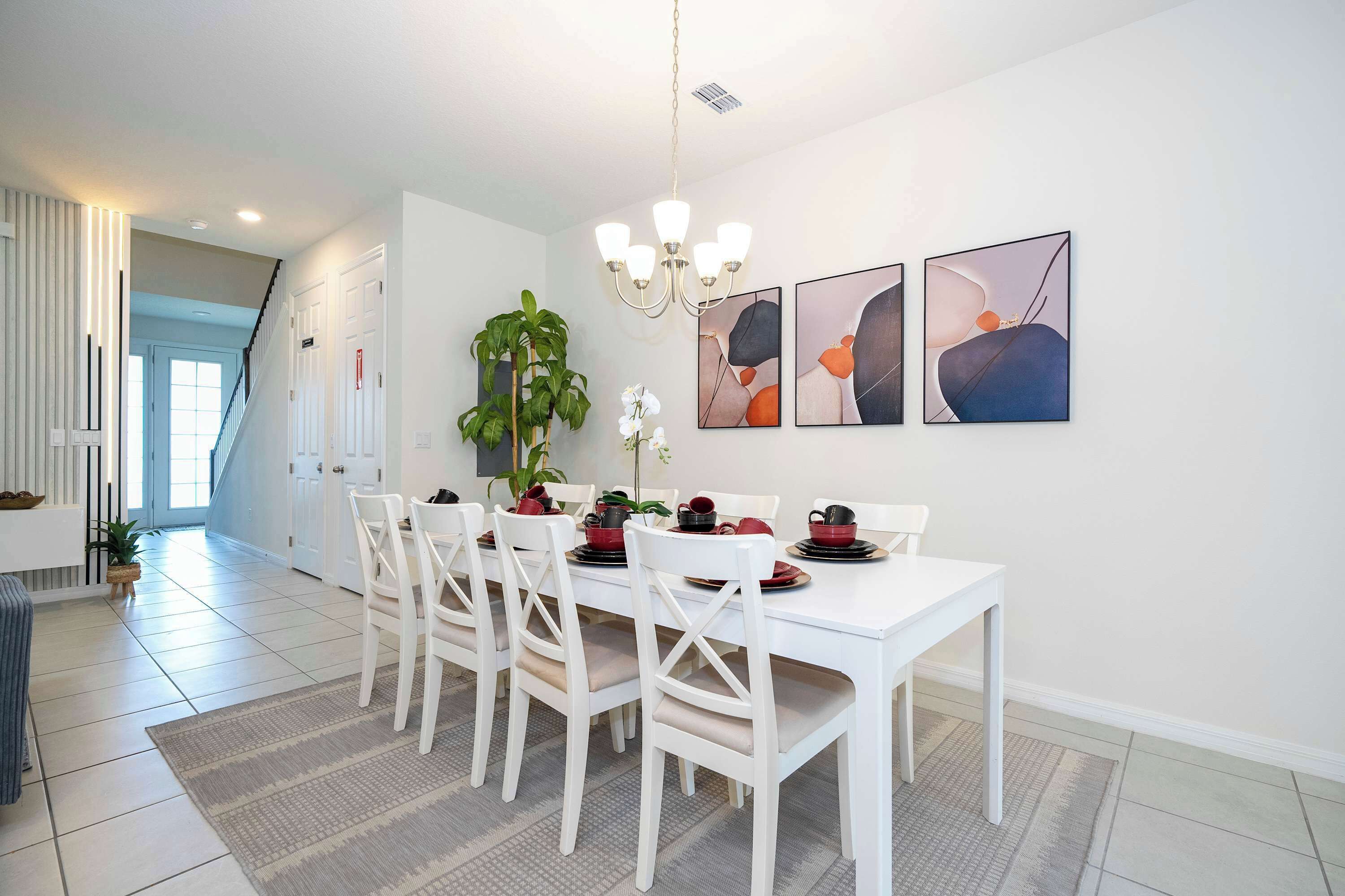 A dining area with a white table, chairs, and a chandelier. There are abstract paintings on the wall and a potted plant.