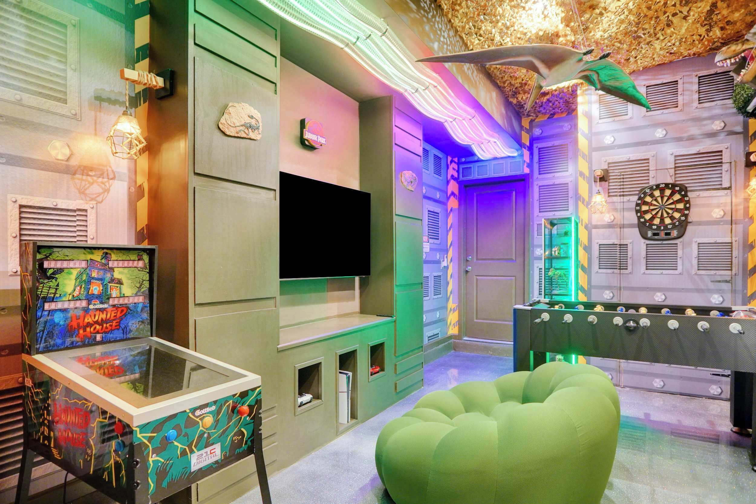 A room with a foosball table, a dartboard, a television, and a pinball machine.