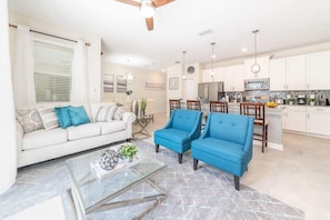 A modern living room with a sofa, two blue armchairs, a glass coffee table, and a kitchen area with white cabinets and a dining set. - Chic, Spacious With Private Pool, Ideal Location (Kissimmee)
