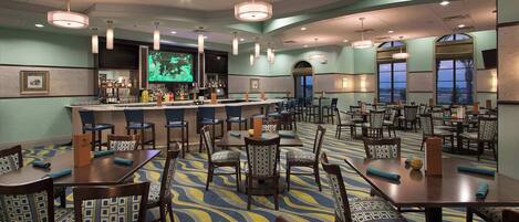 A hotel dining area with multiple tables and chairs, a bar, and large windows.