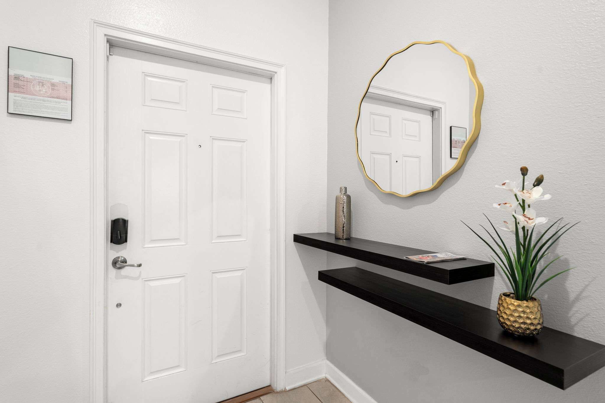 Una puerta blanca con un ojo de cerradura, un espejo fijado a la pared con marco dorado, y dos estantes negros flotantes con una planta y un objeto decorativo.
