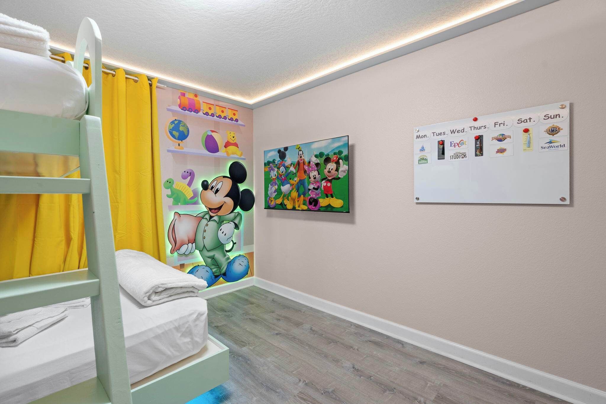 Een kinderkamer met stapelbedden, een muurschildering van Mickey Mouse, een agenda en een plank vol speelgoed.