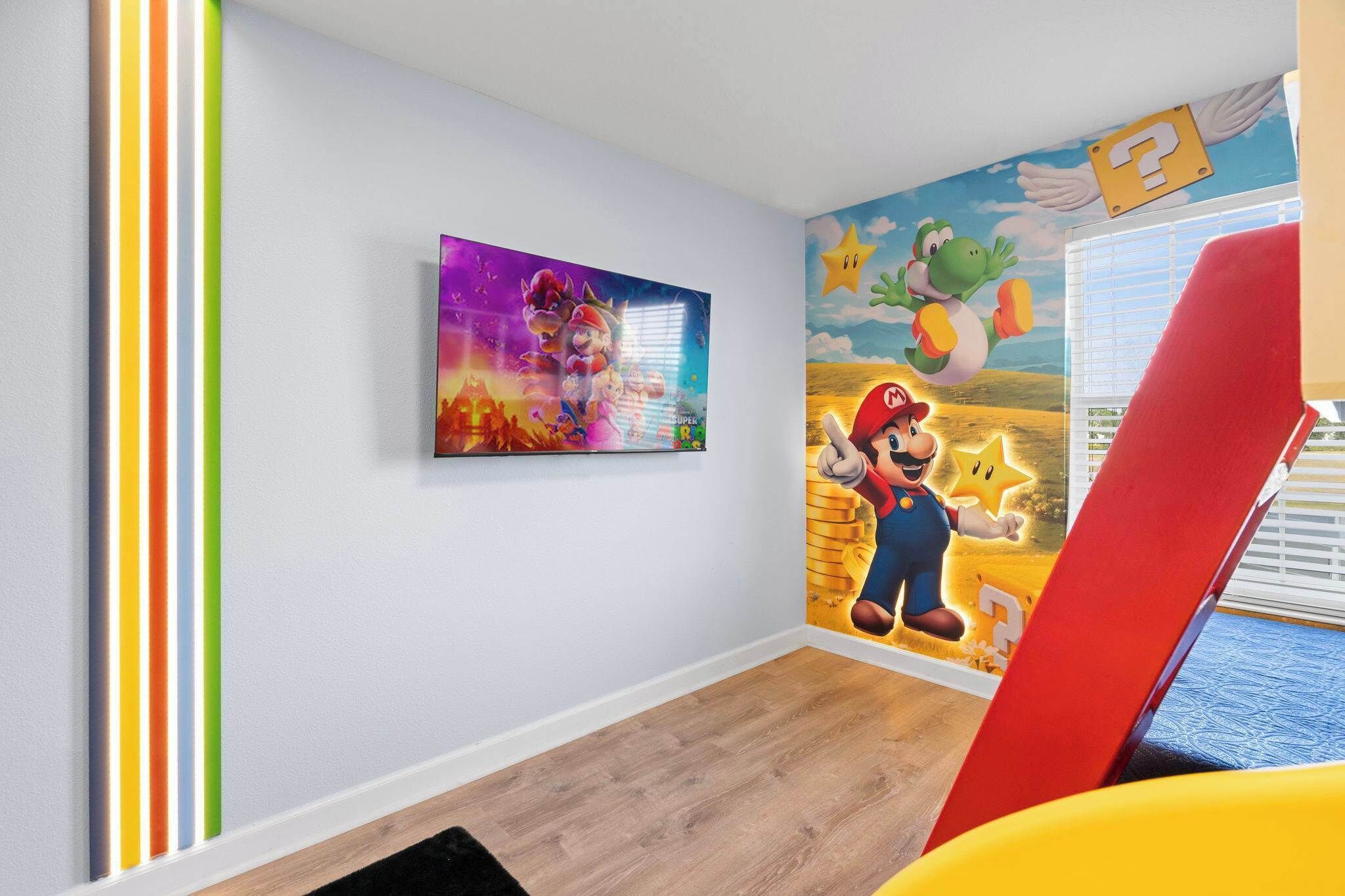 A room with a Mario-themed mural, a framed picture on the wall, and a window with blinds.