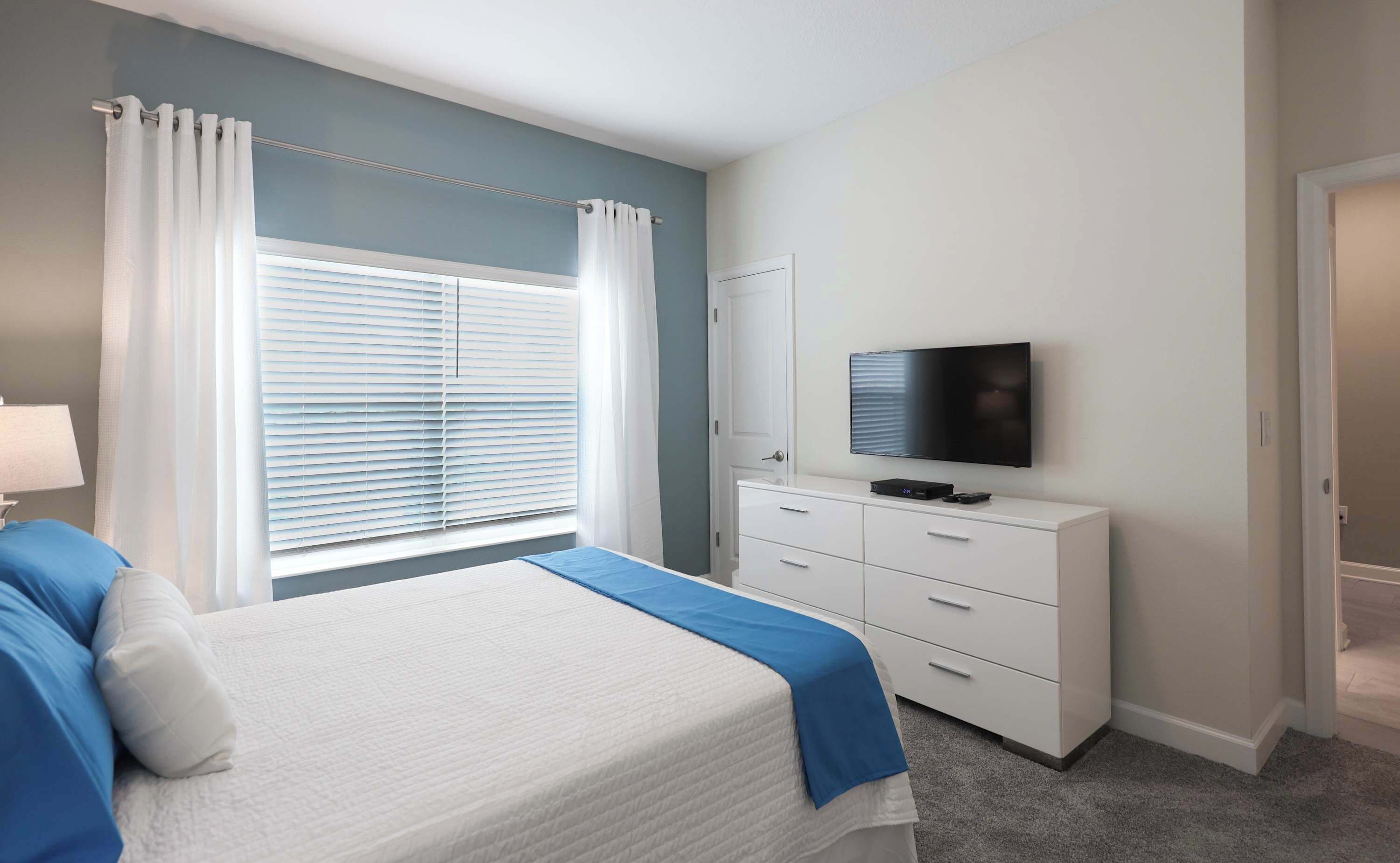 Quarto com cama, penteadeira, televisão e janela com cortinas.