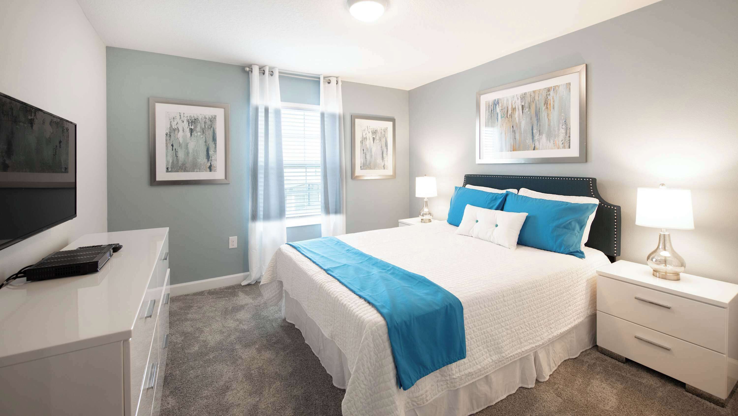 A bedroom with a bed, bedside tables, a desk, a TV, and artwork on the walls.