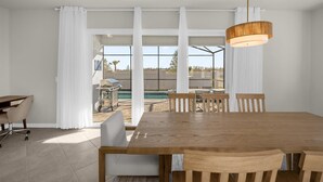 A spacious dining area with a large wooden table and chairs, white curtains, and a view of a pool and outdoor seating. - Elegant 6BR Home Perfect for Families (Clermont)