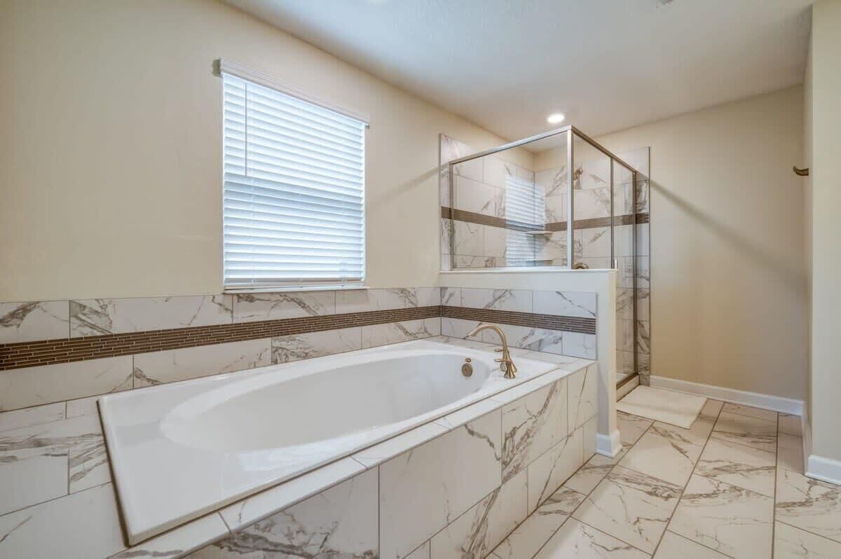 A bathroom with a large bathtub, a window with blinds, and a glass-enclosed shower area.