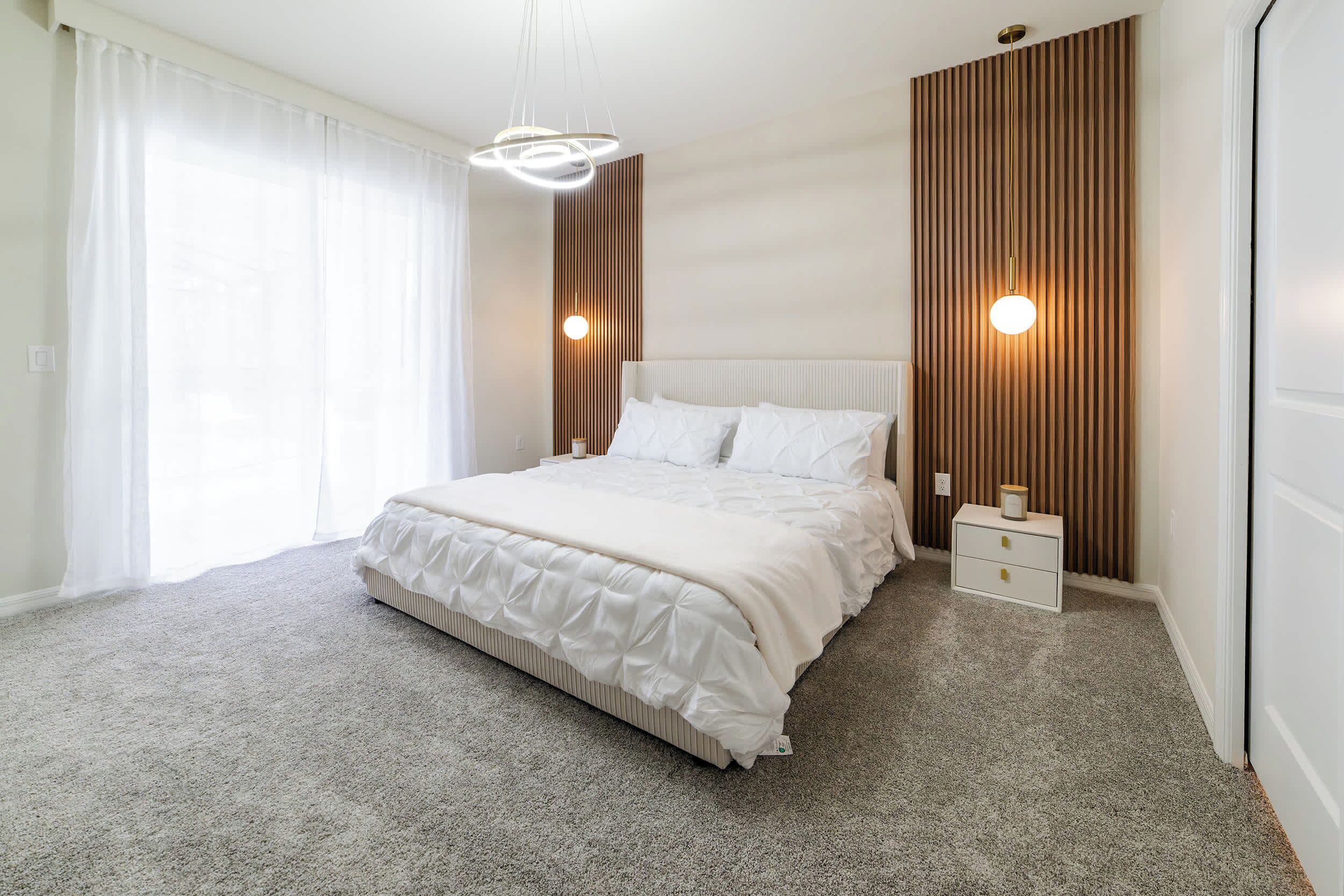 Ein modernes Schlafzimmer mit einem großen Bett, einem Nachttisch, einer Wandleuchte und einem Fenster mit durchsichtigen Vorhängen.