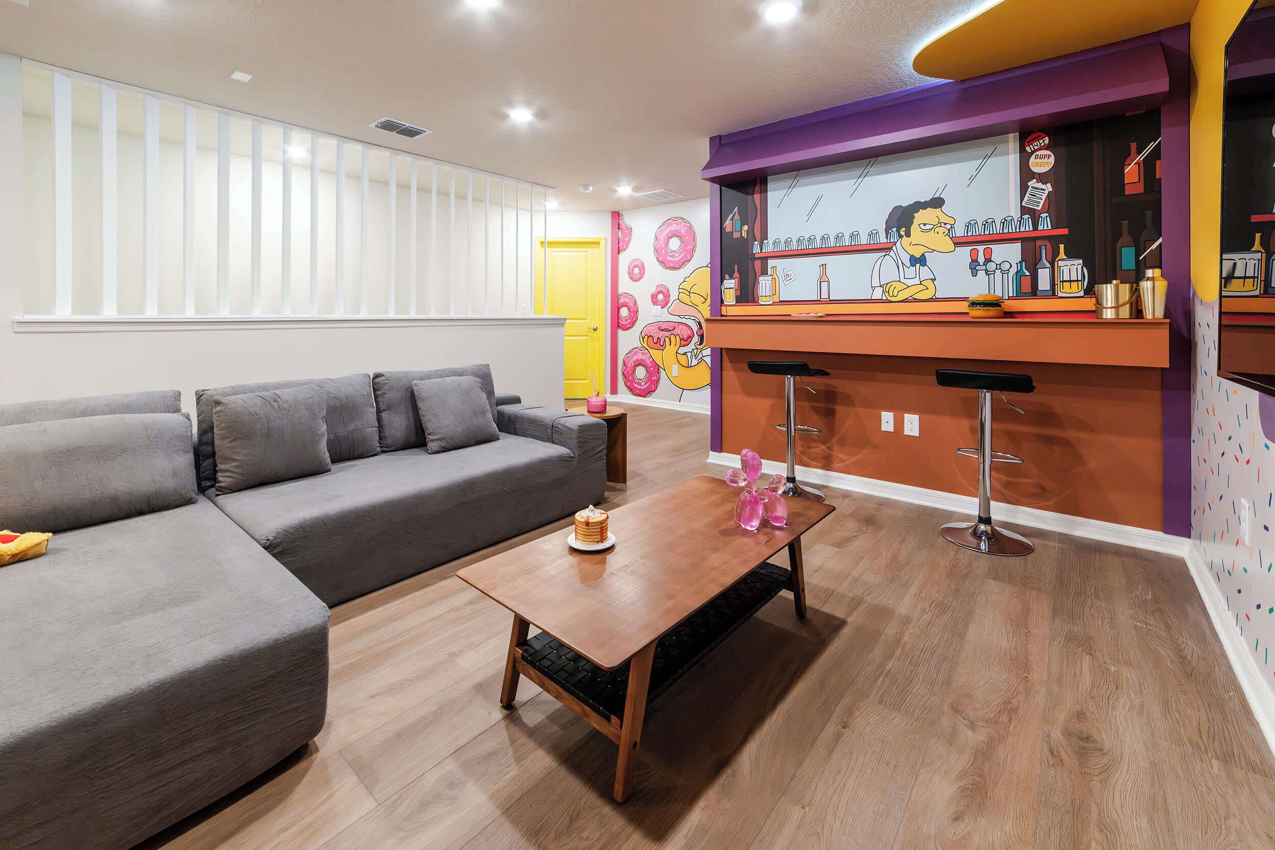 A modern living room with a grey sectional sofa, a wooden coffee table, and a bar area with a cartoon character mural.