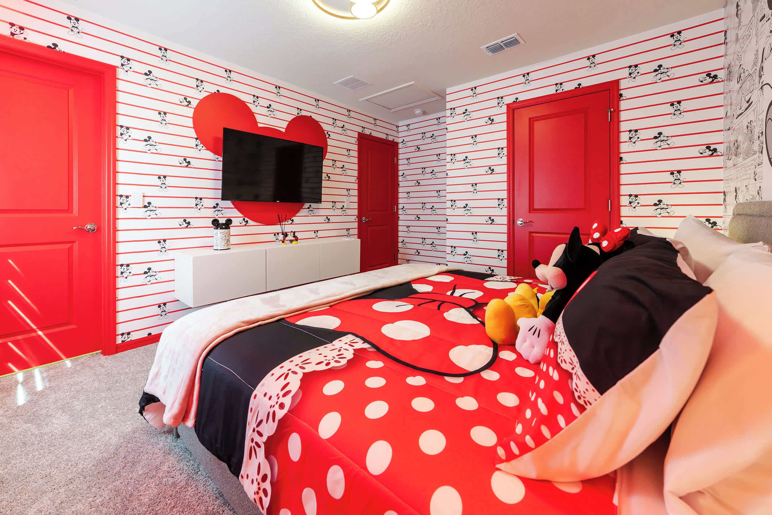 A bedroom with a red and white Mickey Mouse themed wallpaper, a heart-shaped TV mount, and a bed with a polka dot blanket.